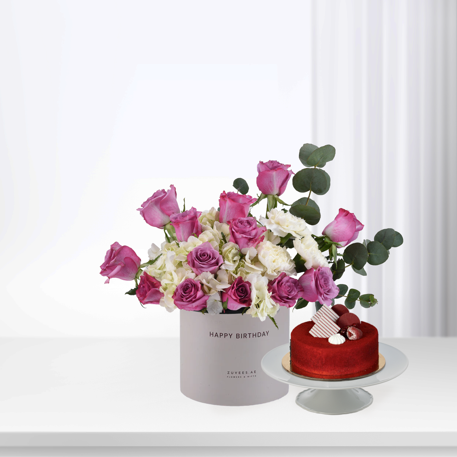 Pink roses, white hydrangeas, and eucalyptus in gray "Happy Birthday" box beside red velvet cake.
