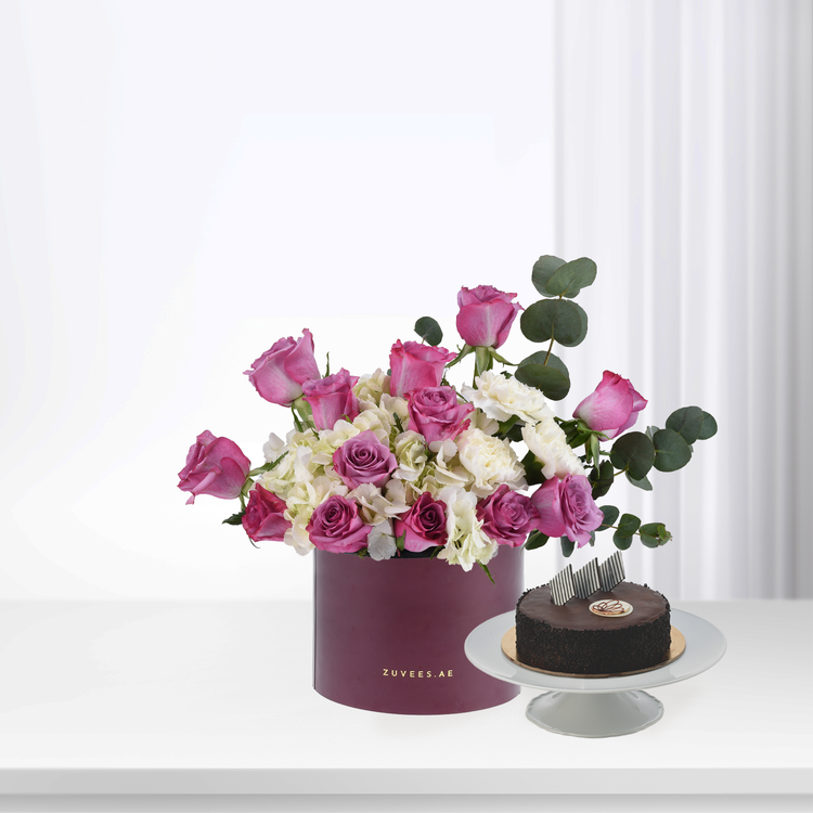 Pink roses, white hydrangeas, and eucalyptus in burgundy box beside chocolate cake.