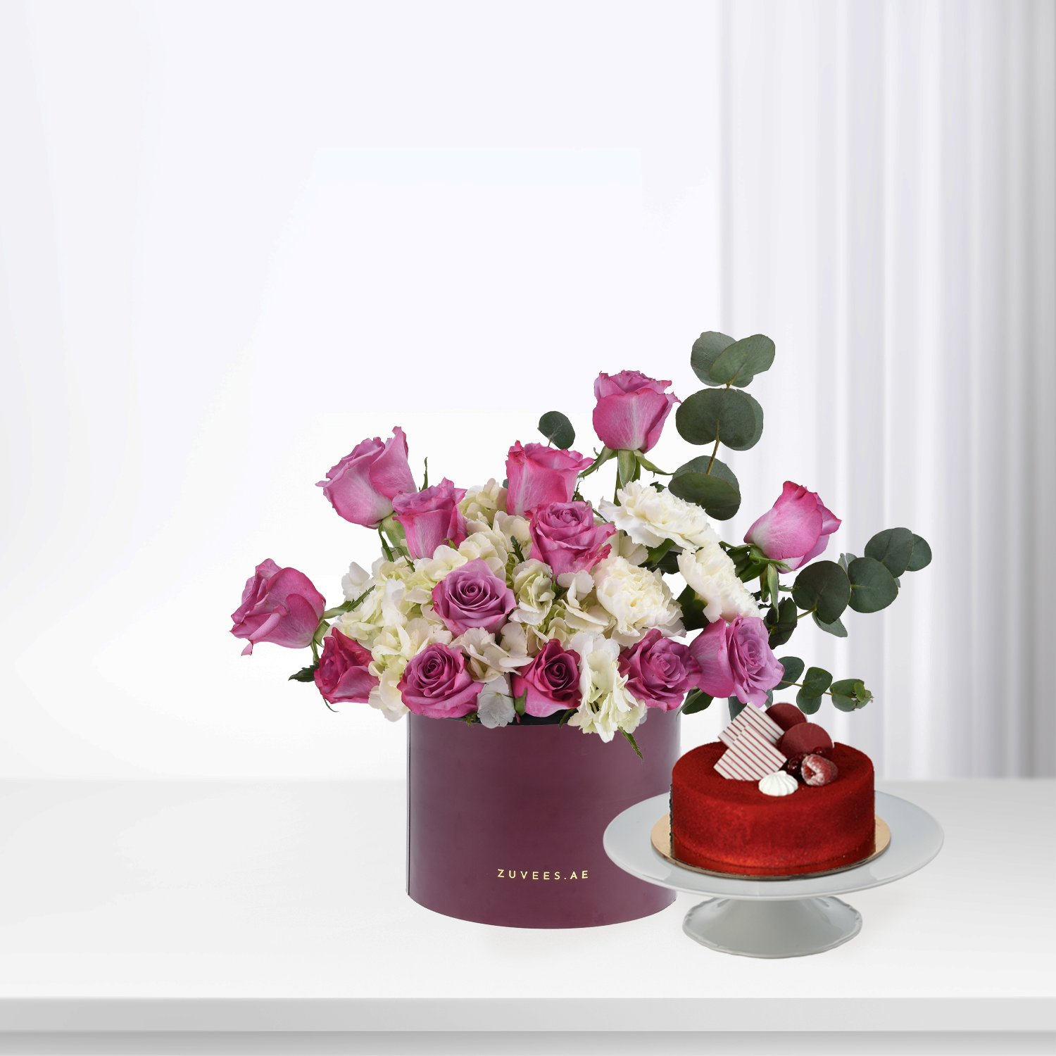 Elegant pink roses and white blooms in burgundy box beside red velvet cake.