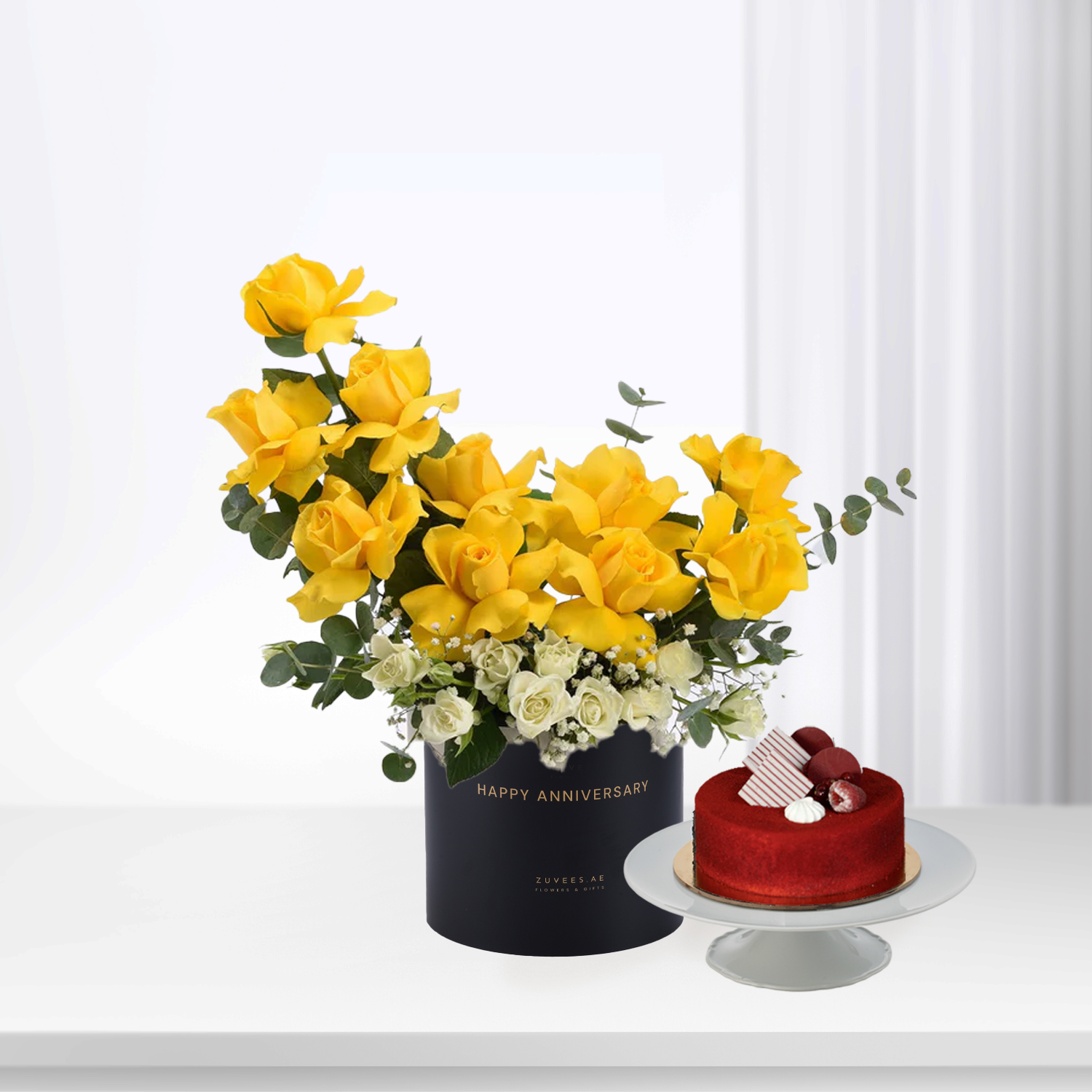 Vibrant yellow roses and white blooms in black "Happy Anniversary" box beside red velvet cake.