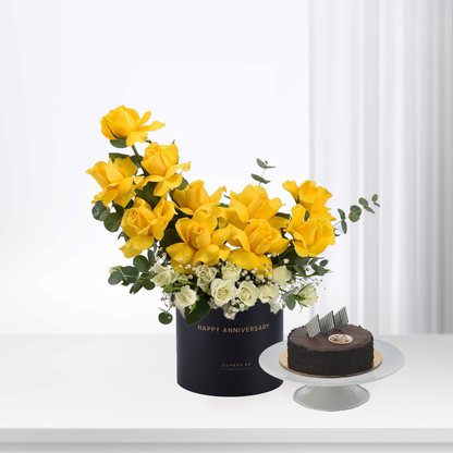 Vibrant yellow roses and white blooms in black "Happy Anniversary" box beside chocolate cake.