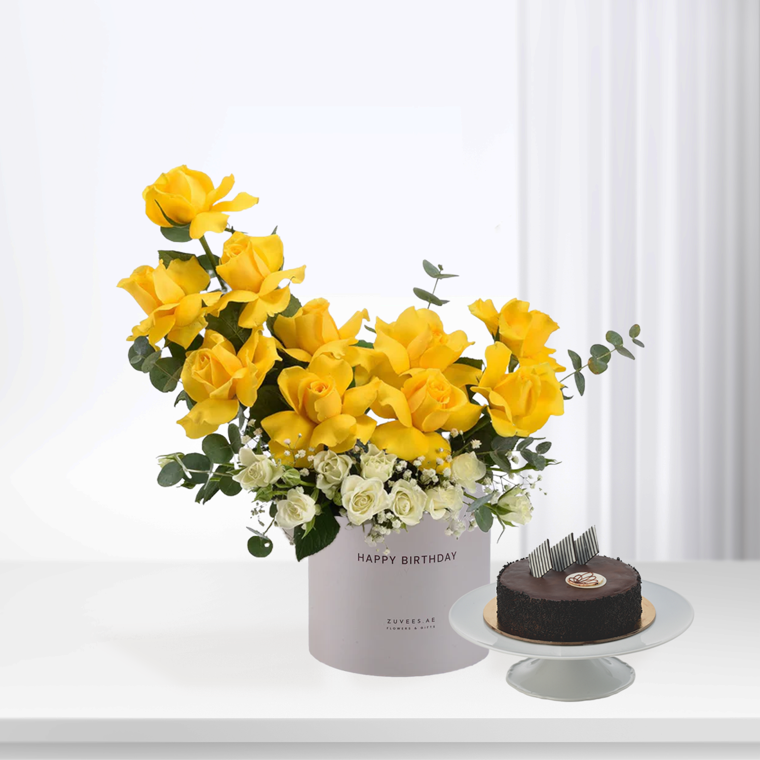 Vibrant yellow roses with white accents in branded box beside chocolate cake.