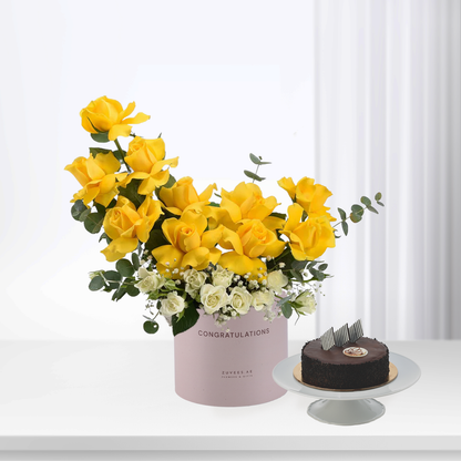 Vibrant yellow roses and white blooms in pink "Congratulations" box beside chocolate cake.