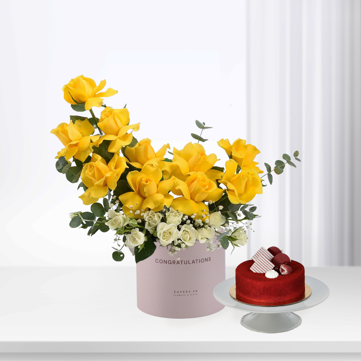 Vibrant yellow roses and white blooms in pink "Congratulations" box beside red velvet cake.