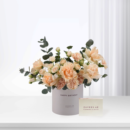 Soft peach roses and carnations in white cylinder with "Happy Birthday" text.