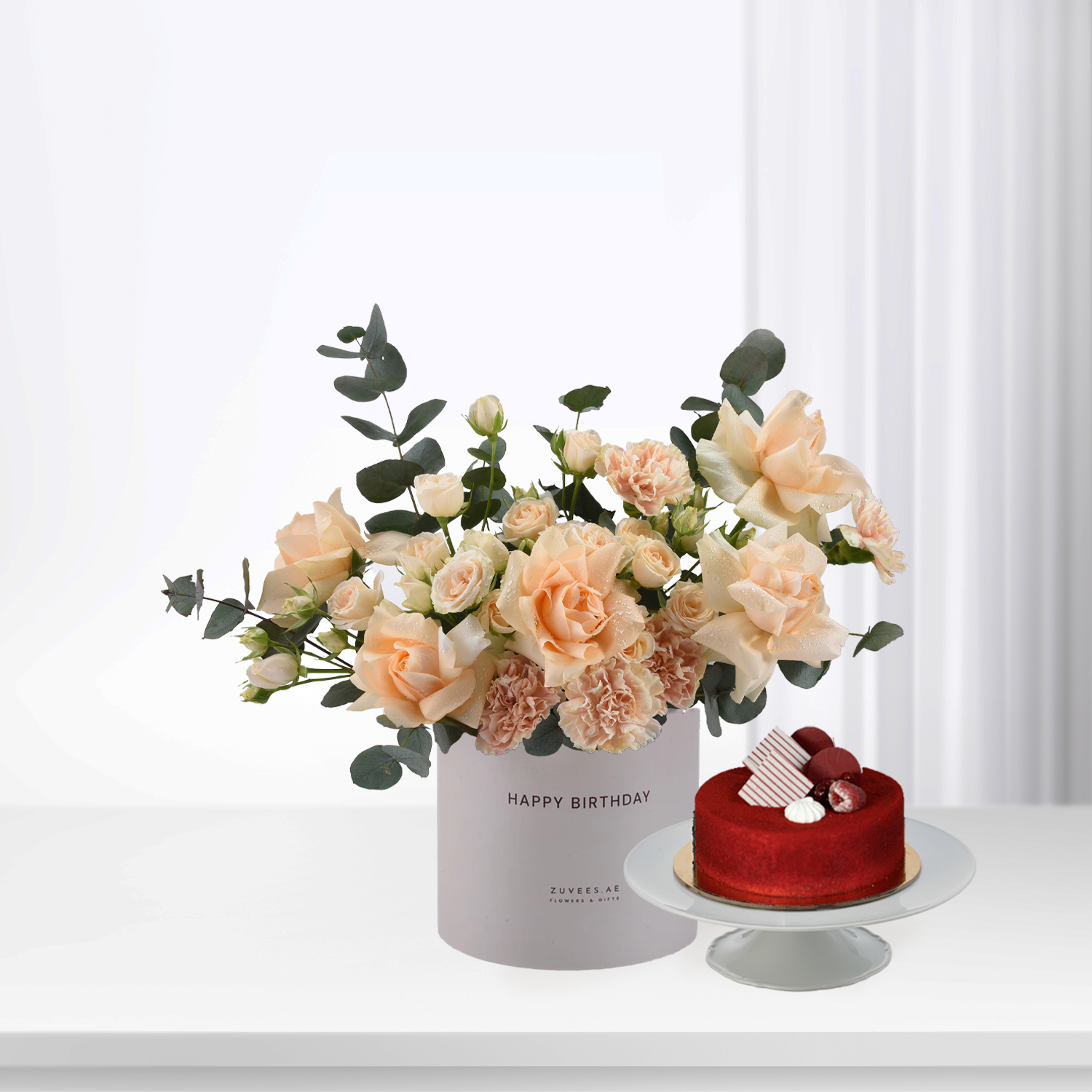 Soft peach roses and eucalyptus in white box beside red velvet cake.
