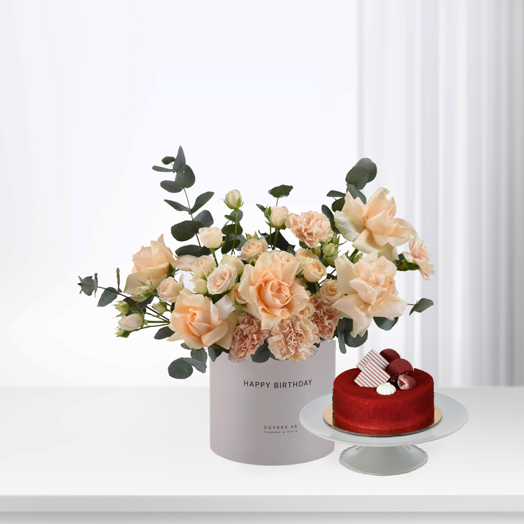 Soft peach roses and eucalyptus in white box beside red velvet cake.