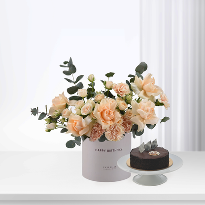 Soft peach roses and carnations in white box beside chocolate cake.