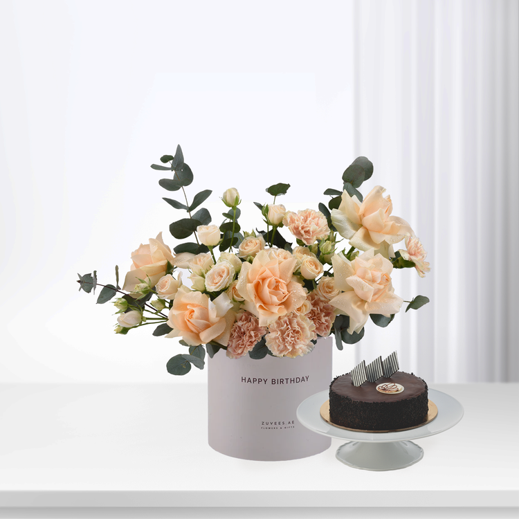 Soft peach roses and carnations in white box beside chocolate cake.