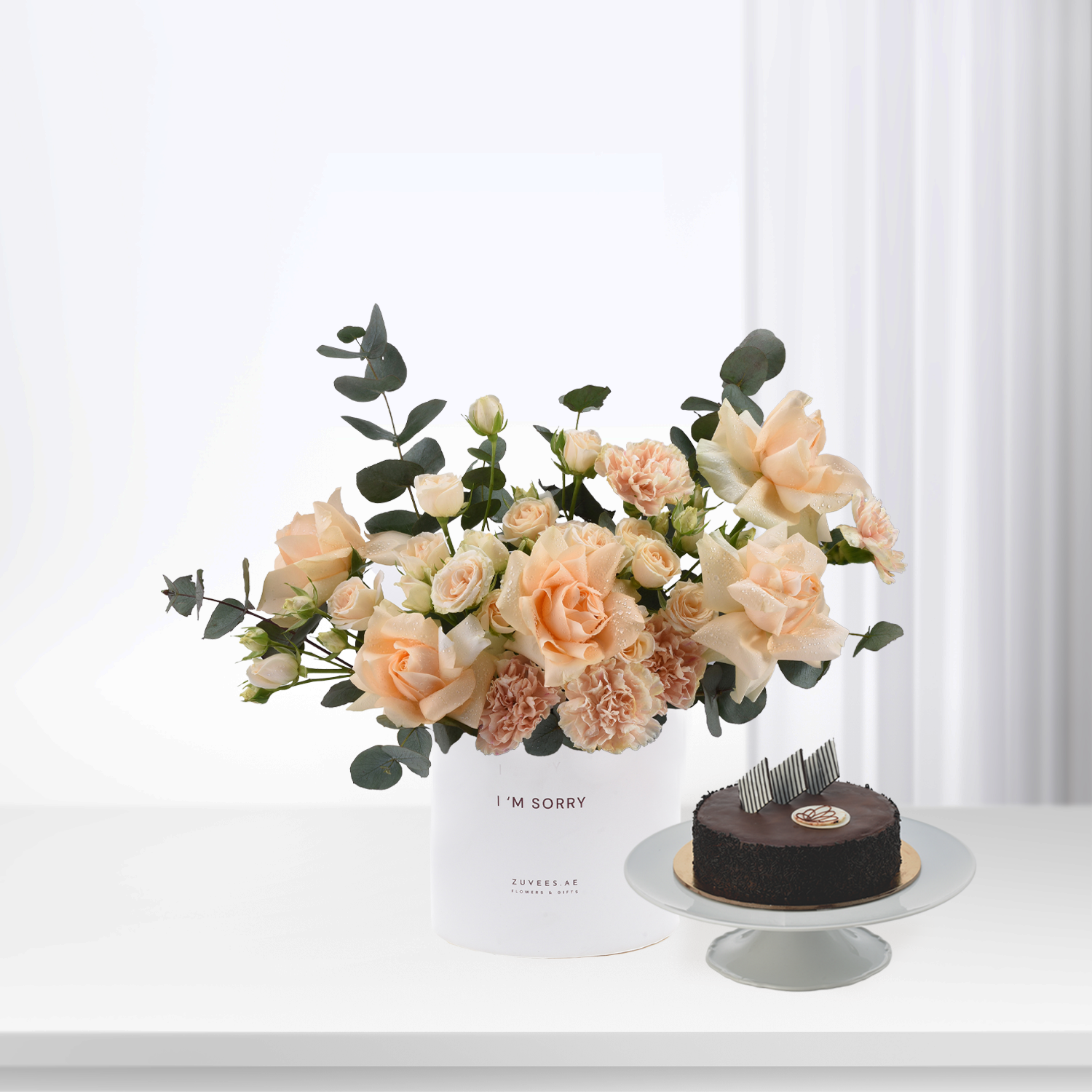 Soft peach roses and eucalyptus in white vase beside chocolate cake.