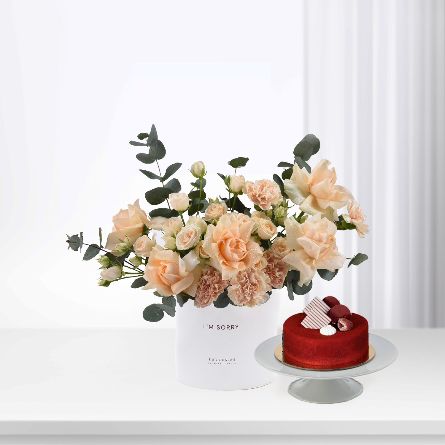 Soft peach roses and eucalyptus in white vase beside red velvet cake.