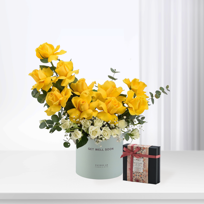 Vibrant yellow roses in mint cylinder with "Get Well Soon" text, beside patterned gift box.