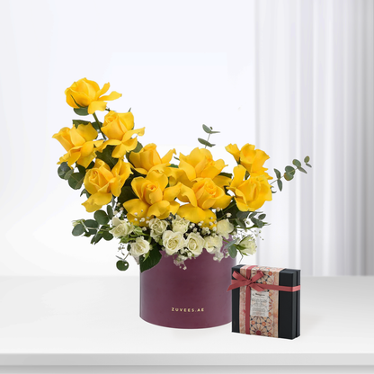 Vibrant yellow roses in burgundy box, accented with white blooms and eucalyptus, beside ornate chocolate gift.