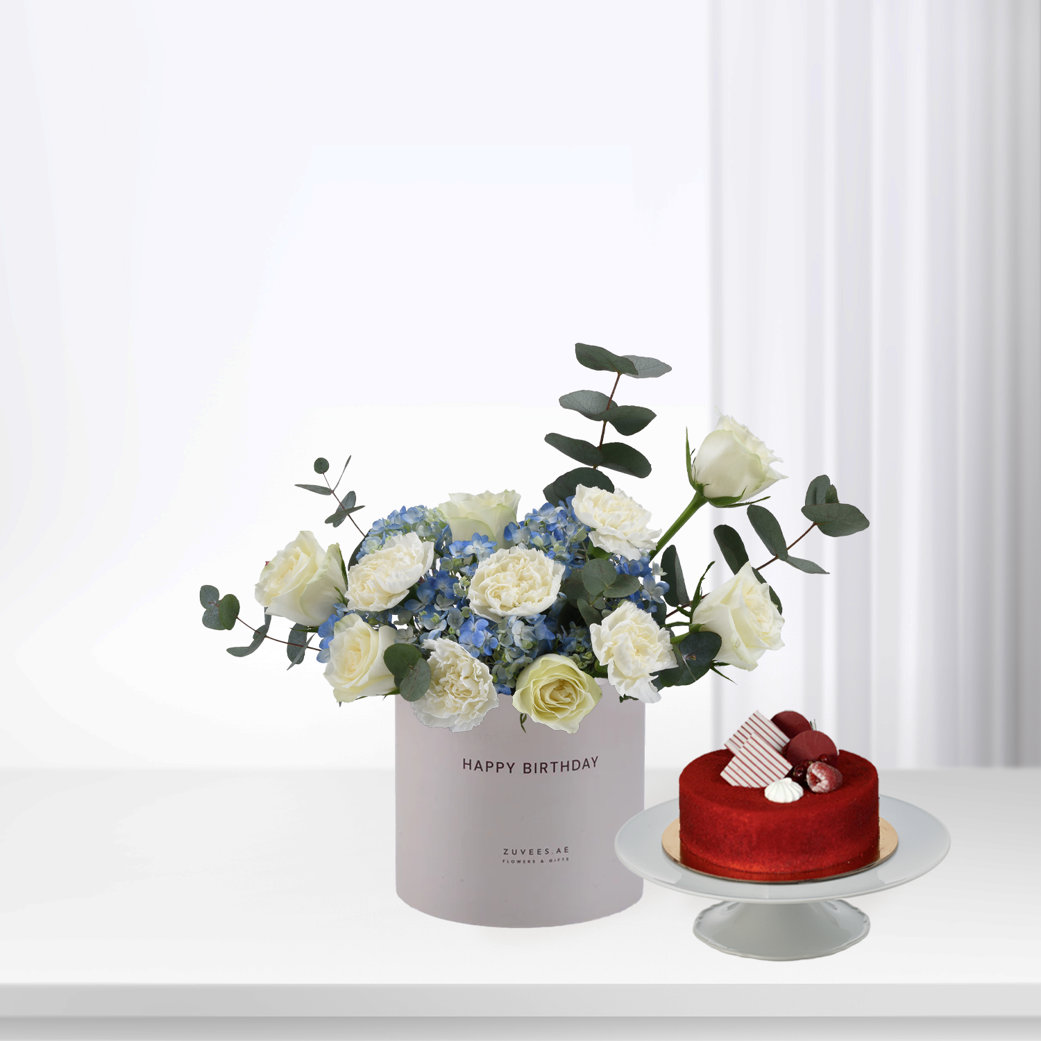 White roses, blue hydrangeas, eucalyptus in "Happy Birthday" box beside red velvet cake.