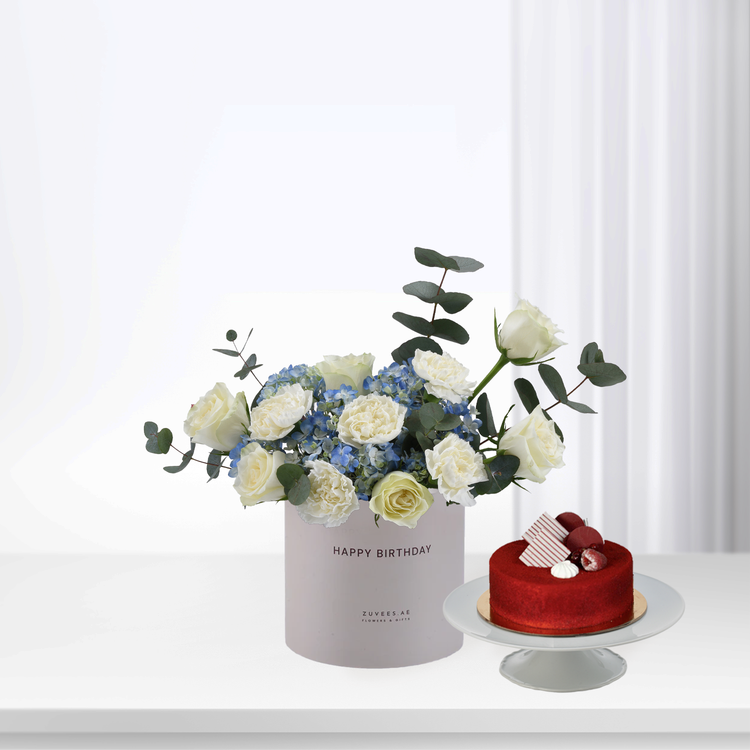 White roses, blue hydrangeas, eucalyptus in "Happy Birthday" box beside red velvet cake.