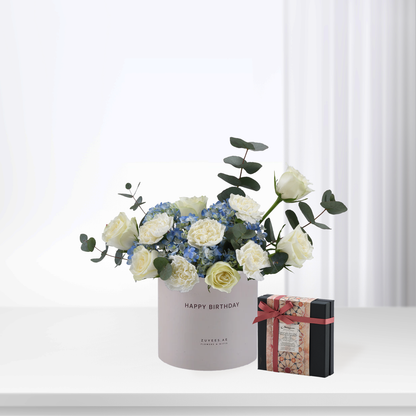 White roses and blue hydrangeas in matte pink "Happy Birthday" box beside patterned gift.