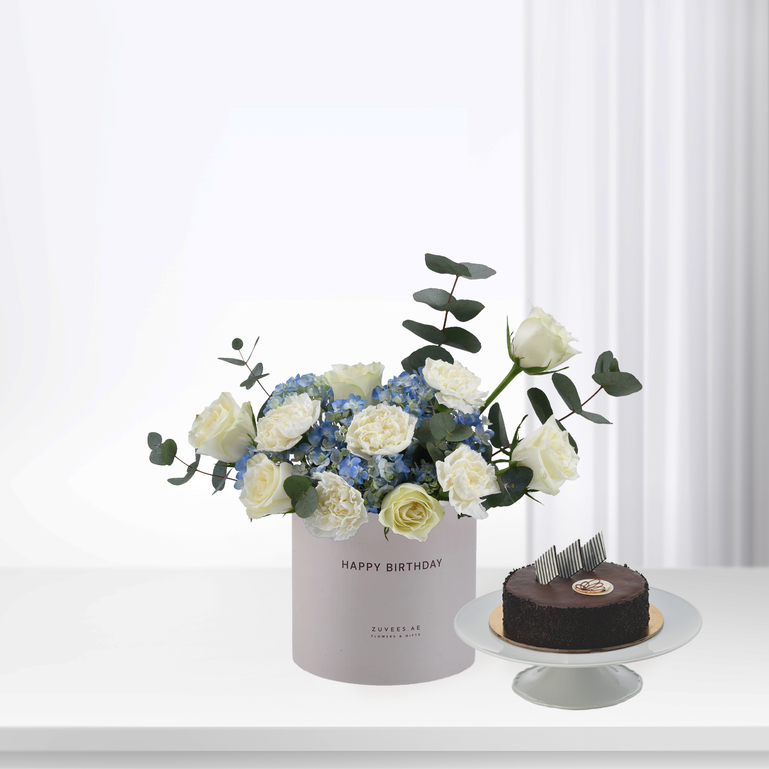 Elegant white roses and blue hydrangeas in pastel box beside chocolate cake.