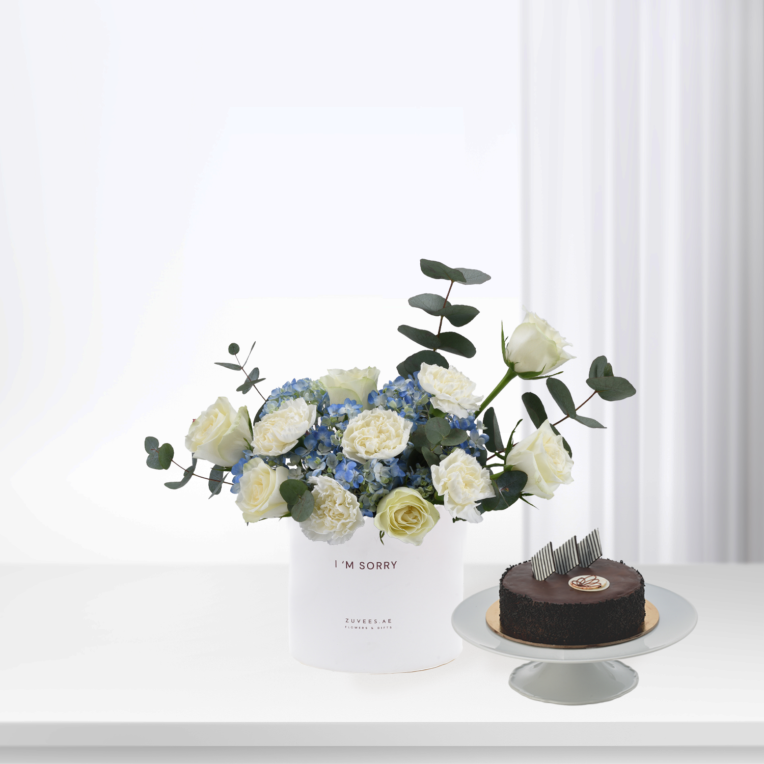 White roses and blue hydrangeas in minimalist vase beside chocolate cake.