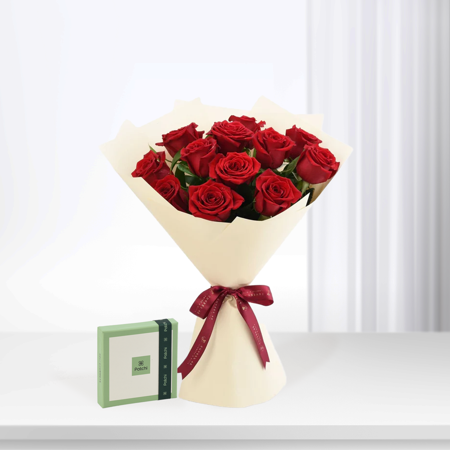 Vibrant red roses in cream paper, tied with burgundy ribbon, beside green Patchi box.