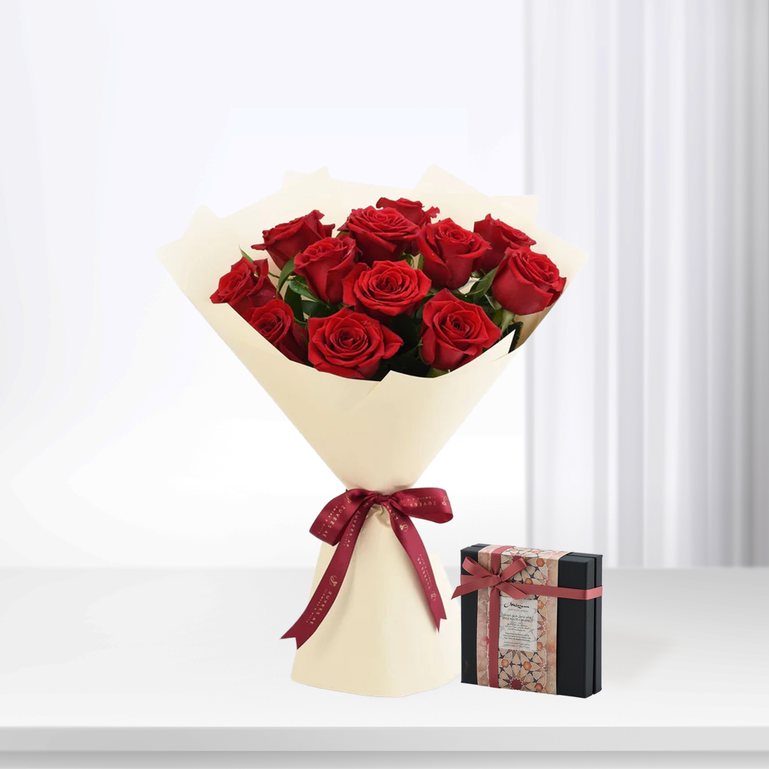 Vibrant red roses in cream wrap with burgundy ribbon, paired with patterned gift box on white surface.
