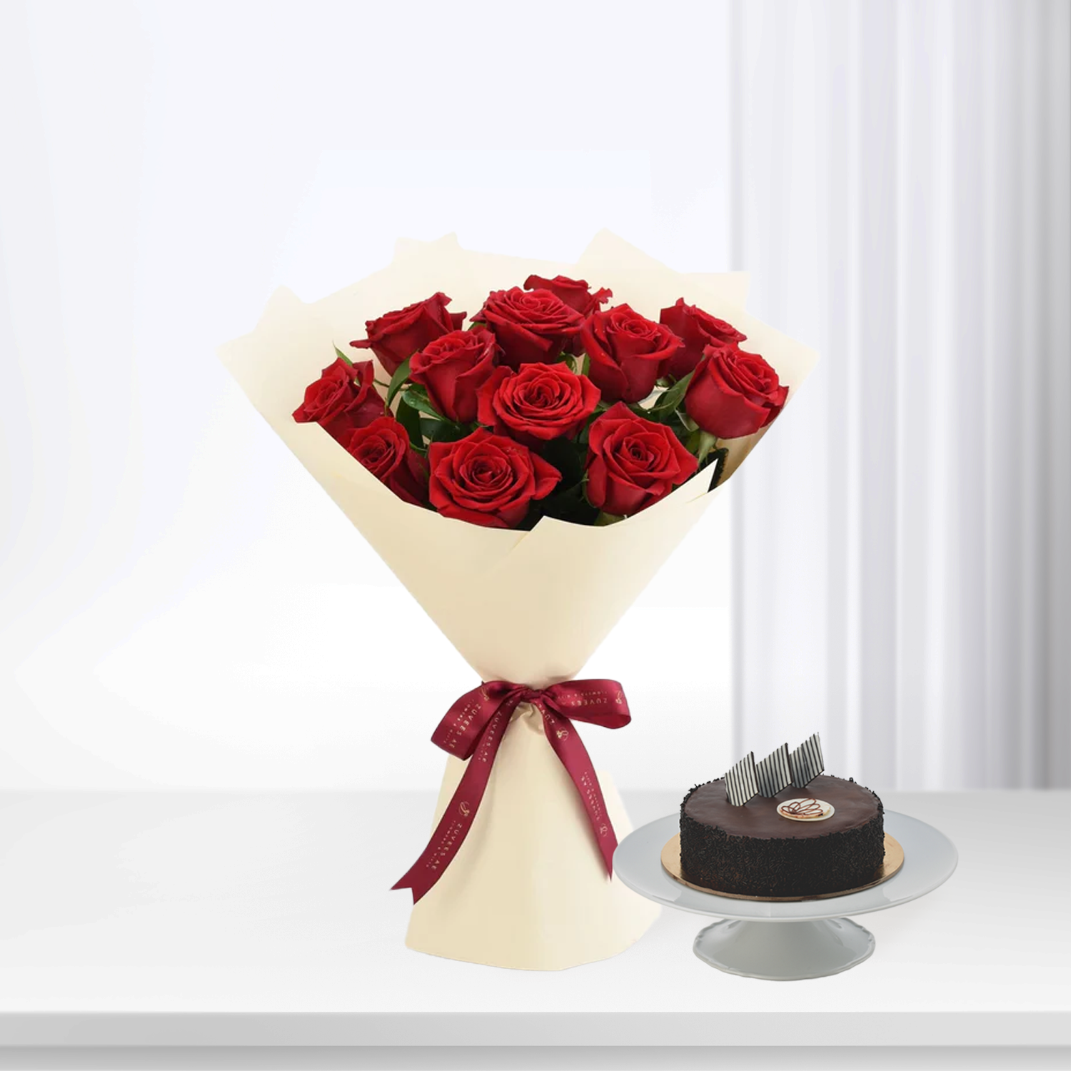Vibrant red roses in cream wrap with burgundy ribbon, beside chocolate cake on stand.