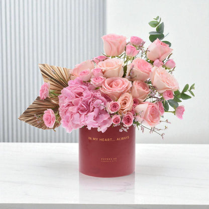 Soft pink roses and hydrangeas bloom in a matte red vase with gold text.