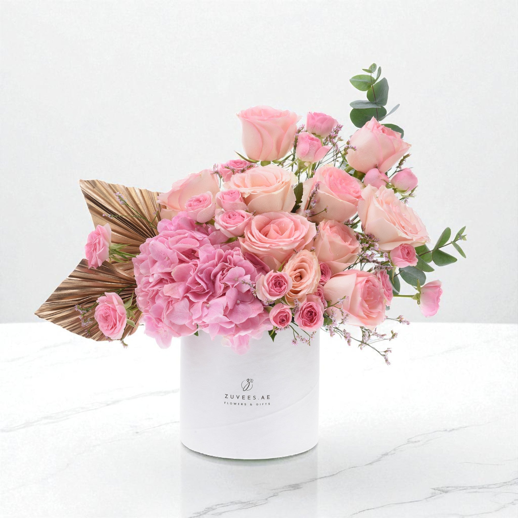 Pink roses, hydrangeas, gold palm leaves in white Zuvees box on marble.