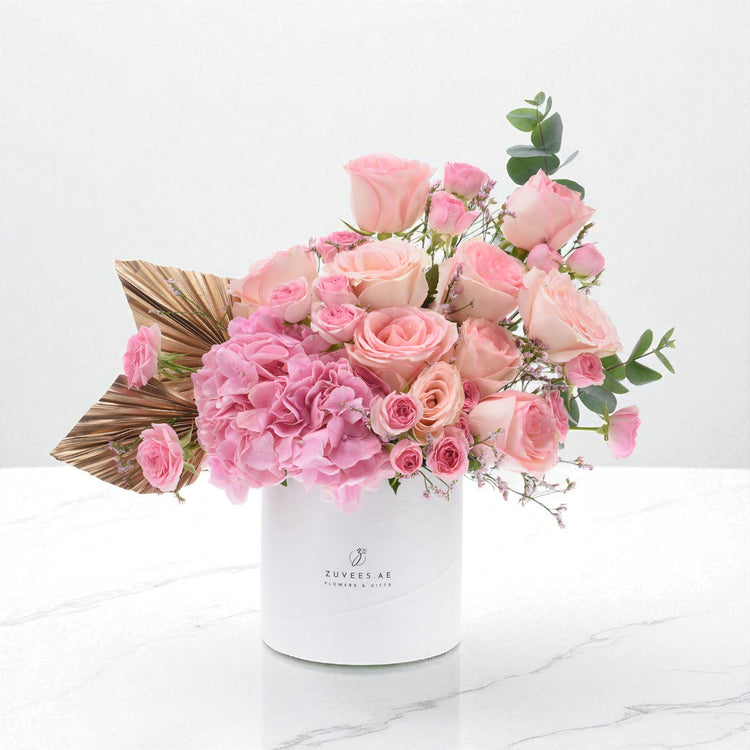 Pink roses, hydrangeas, gold palm leaves in white Zuvees box on marble.