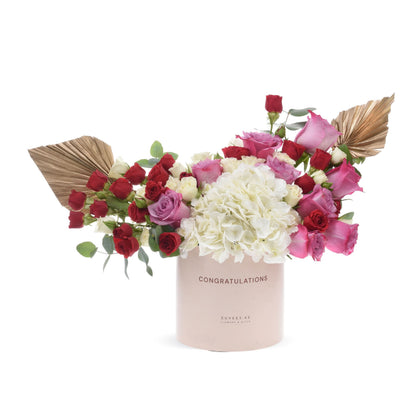 Pink box holds vibrant red, pink, and white roses with gold fan accents.