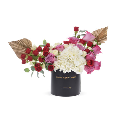 Elegant anniversary bouquet in black box with gold text, red, pink, and white roses, white hydrangeas, and gold fan leaves.