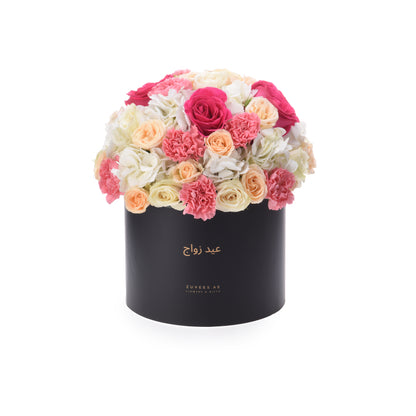 Black cylindrical box overflowing with vibrant pink, peach, and white roses and carnations.