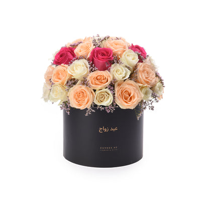 Black box holds vibrant peach, pink, and cream roses with delicate purple filler blooms.