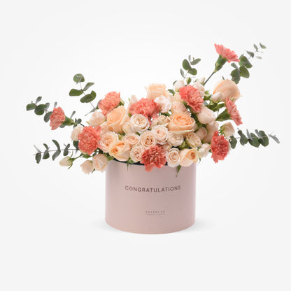 Soft pink box with "CONGRATULATIONS" text, holding peach roses and carnations with eucalyptus.